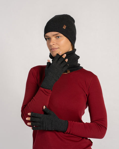 Side-profile of a woman in a dark-gray beanie and gaiter, wearing dark-gray fingerless gloves and looking at her raised hand.