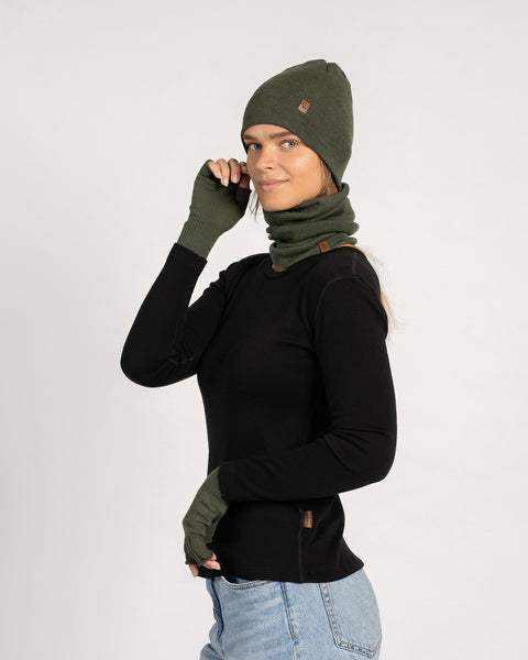 Woman in a dark-green beanie and matching gaiter and gloves, smiling in a three-quarter pose with one hand near her head.