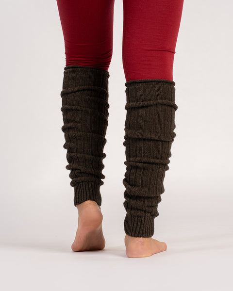A view from behind of a person standing barefoot, wearing brown, ribbed-knit leg warmers scrunched down over red leggings against a solid light gray background.
