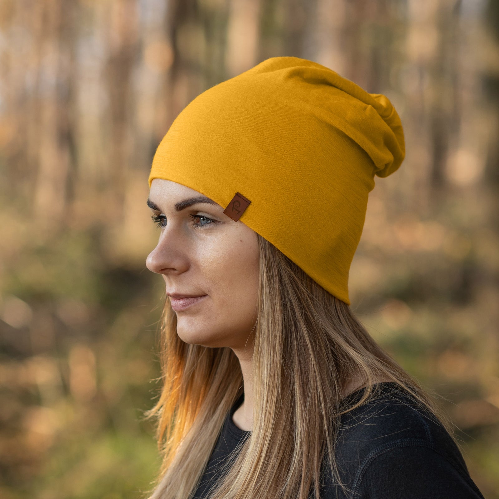 A close-up shot of a spicy yellow merino wool menique beanie hat is presented against a stark white background. The beanie has a slightly textured appearance and features a small, brown rectangular tag sewn onto its folded brim. The hat is softly draped and shows gentle wrinkles in the fabric.