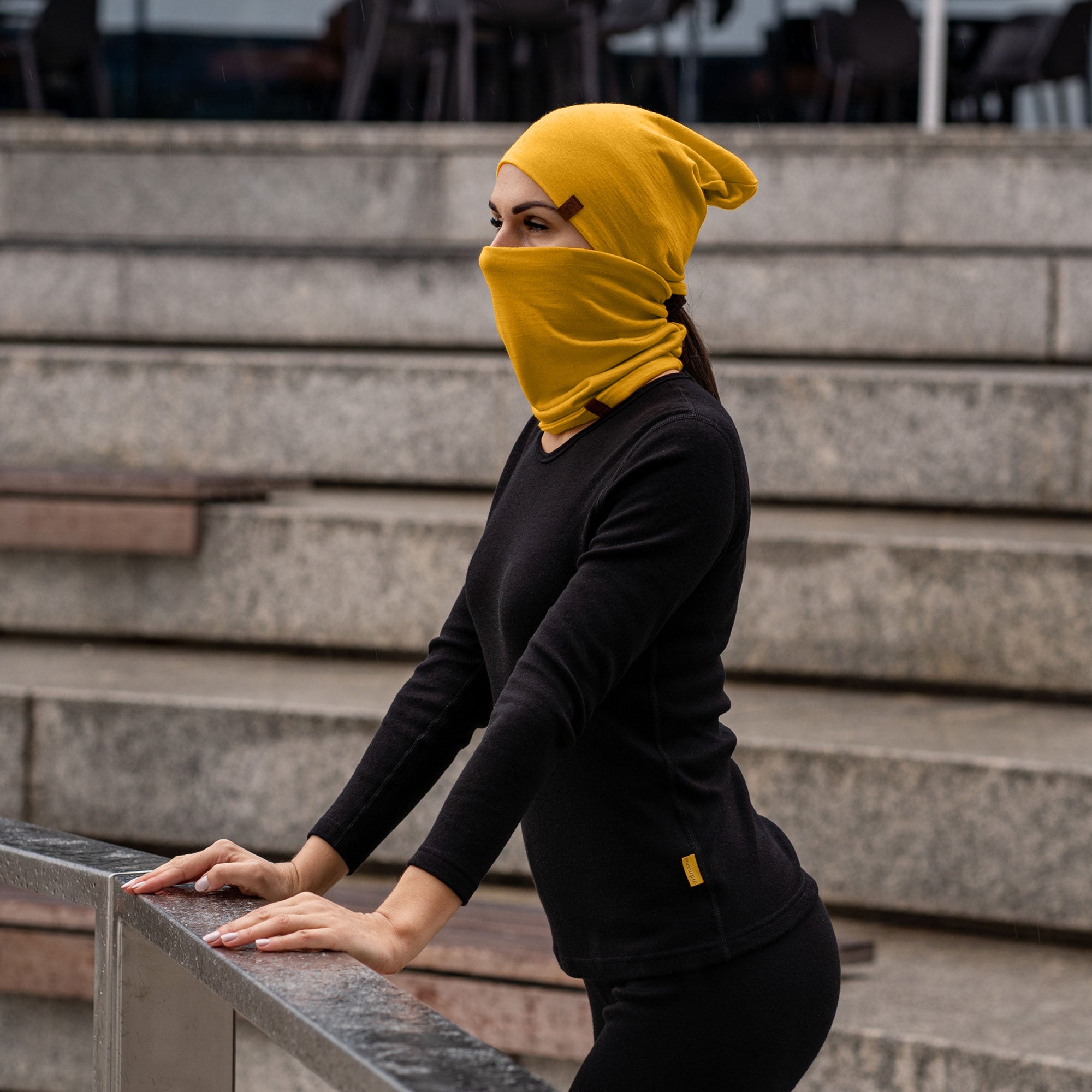 A fair-skinned individual with light-colored eyes is wearing a spicy yellow merino wool beanie that covers their head and a matching neck gaiter pulled up over their mouth and nose, leaving only their eyes visible. They are wearing a black top, and the background is plain white.