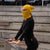 A fair-skinned individual with light-colored eyes is wearing a spicy yellow merino wool beanie that covers their head and a matching neck gaiter pulled up over their mouth and nose, leaving only their eyes visible. They are wearing a black top, and the background is plain white.