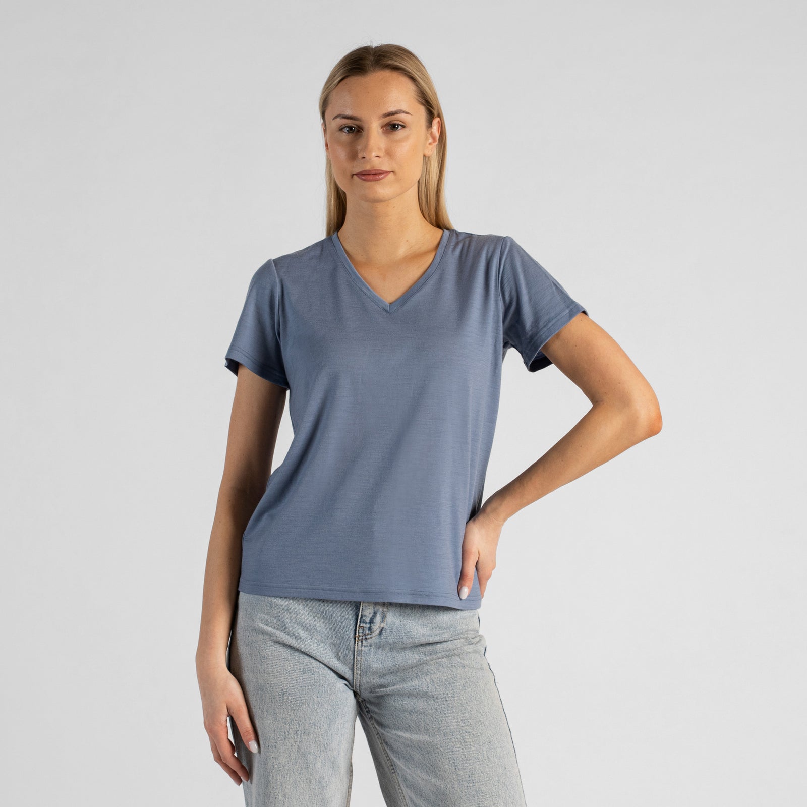 A studio portrait of a blonde woman sitting on a stool and looking directly at the camera. She is leaning back slightly, with her hands resting on her lap. She is wearing a regular fit, dusty blue short-sleeved v-neck t-shirt and light-wash blue jeans against a plain light grey background.