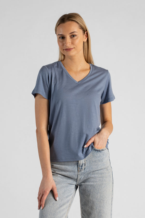 A studio portrait of a blonde woman standing and looking directly at the camera with a subtle smile. She is wearing a dusty blue short-sleeved V-neck t-shirt and light wash denim jeans, with one hand casually resting in her front pocket against a plain light grey background.