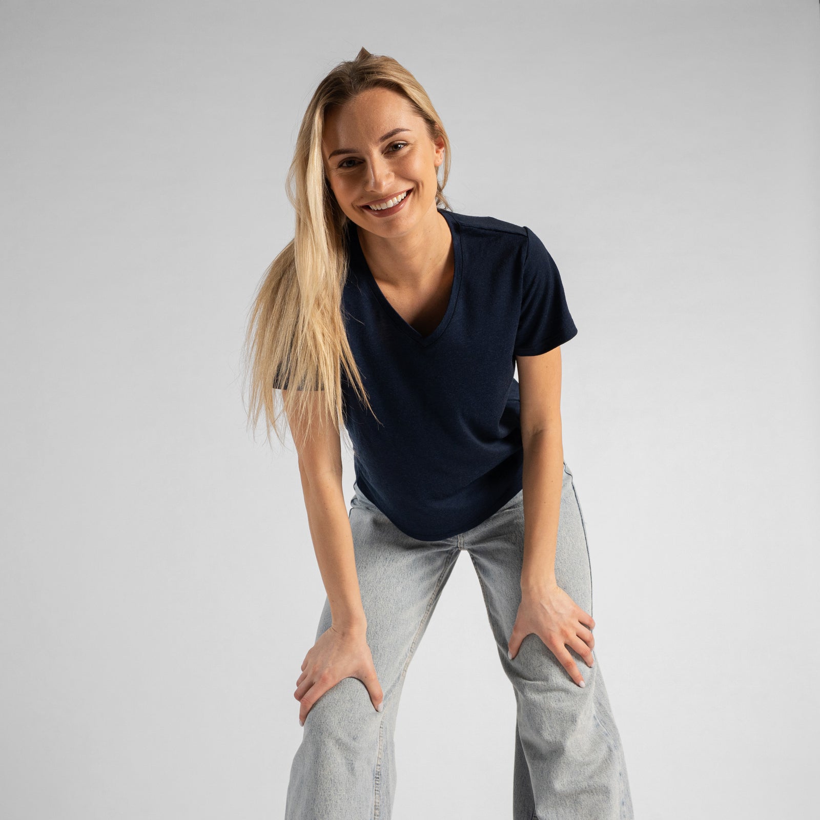 A woman standing next to a textured, rusty metal wall, smiling and looking away from the camera. She is wearing a navy blue v-neck t-shirt and blue jeans, with her right hand resting gently on the metal structure and her left hand in her pocket.