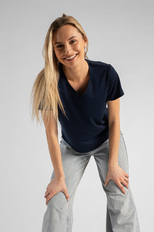 A studio portrait of a blonde woman leaning forward with a big smile, looking directly at the camera. Her hands are resting on her thighs just above her knees. She is wearing a regular fit, dark navy blue short-sleeved v-neck t-shirt and light-wash blue jeans against a plain light grey background.