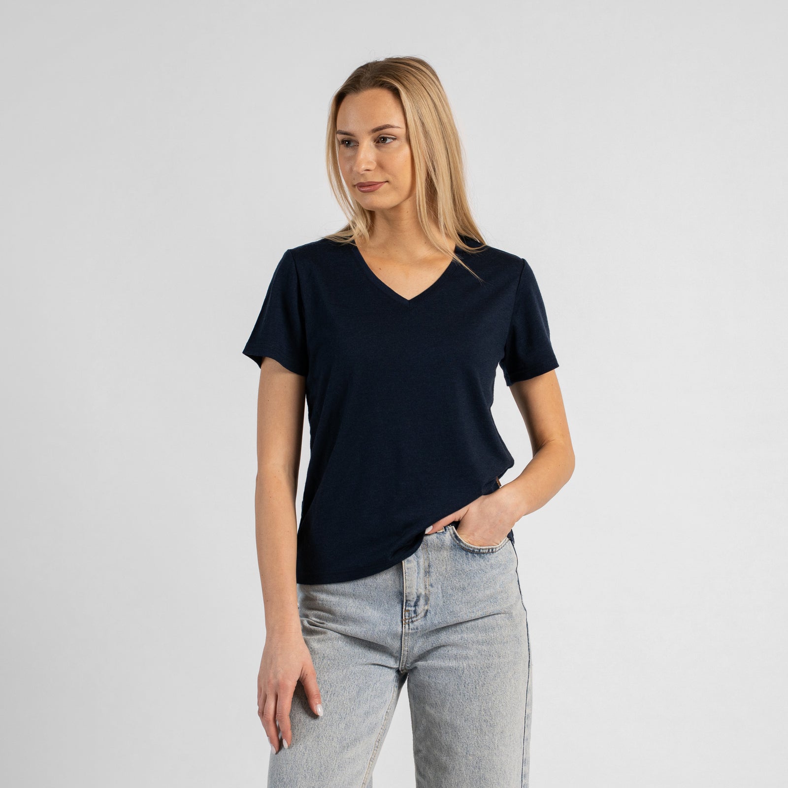A studio portrait of a blonde woman standing and looking directly at the camera with her arms relaxed by her sides. She is wearing a regular fit, dark navy blue short-sleeved v-neck t-shirt and light-wash blue jeans. The background is a solid light grey.