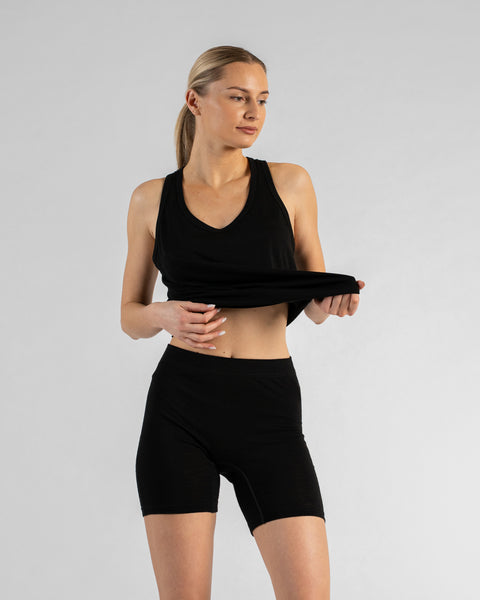 A front view of a blonde woman using one hand to lift the front hem of her black sleeveless top, revealing her midriff and demonstrating the high waistband of the black slip shorts she is wearing.