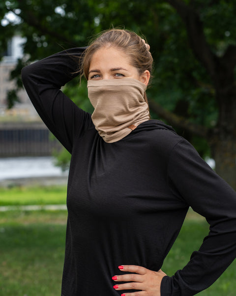 A woman standing outdoors wearing a black long-sleeved top. She has a beige neck gaiter pulled up over her nose and mouth. She is looking at the camera, smiling with her eyes, with one hand on her hip and the other hand resting behind her head, with a background of blurred trees and grass.