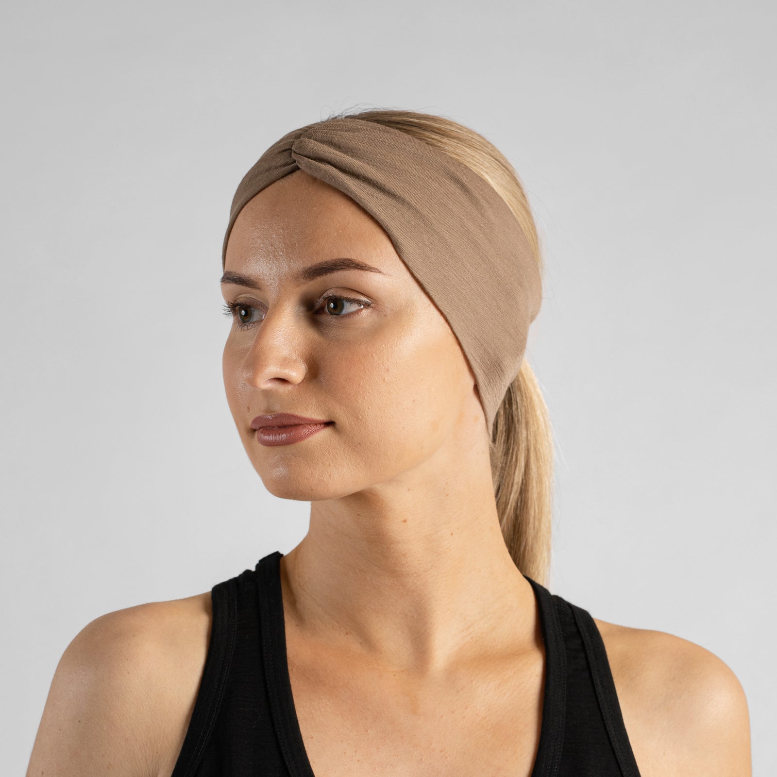 A smiling woman sitting on a blue sports court, wearing a black tank top and a wide textured beige headband.