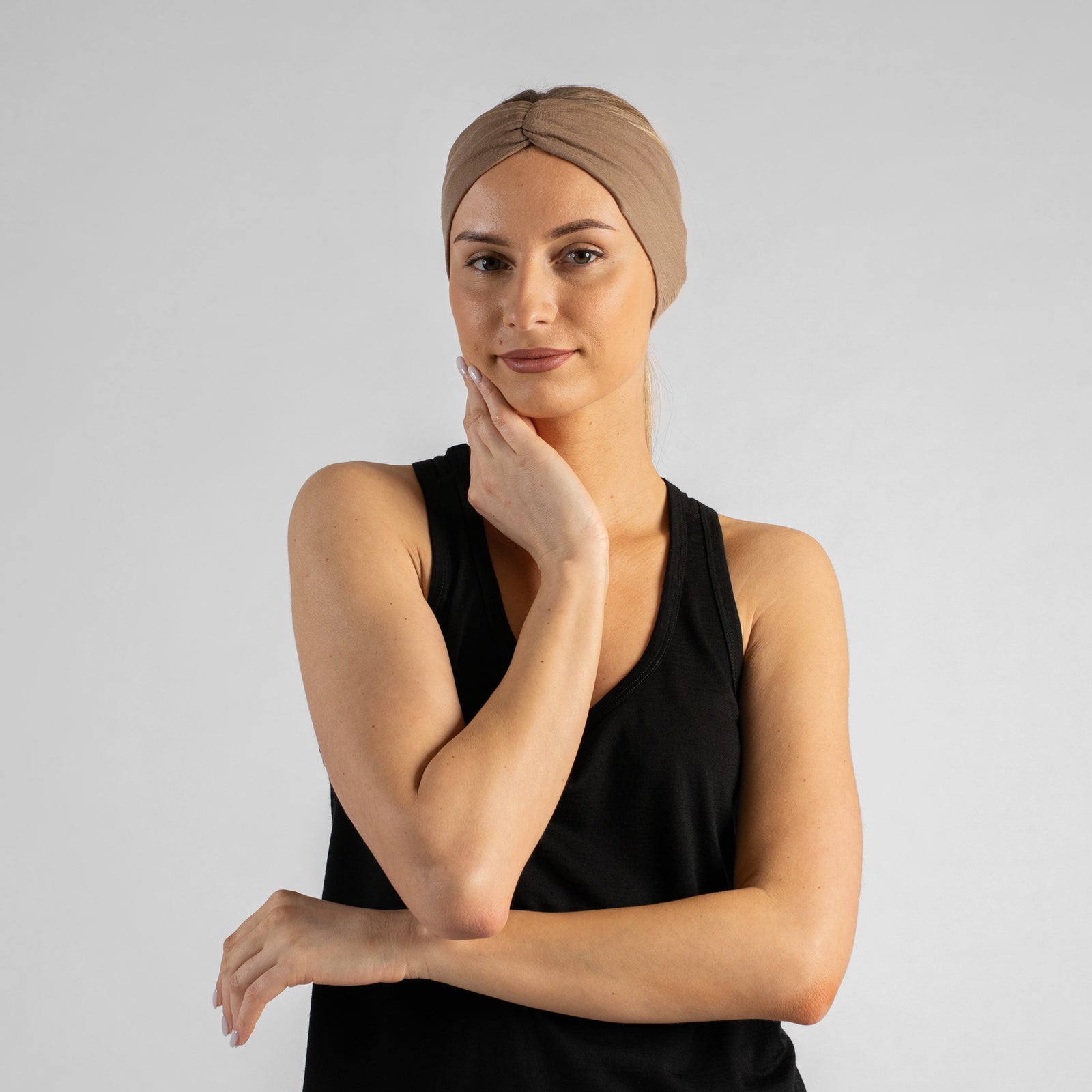 A studio portrait of a blonde woman looking downward with her right hand gently resting on her forehead. She is wearing a black sleeveless top and a beige merino ribbon headband with the twisted ribbon detail centered on her forehead, set against a plain light grey background.