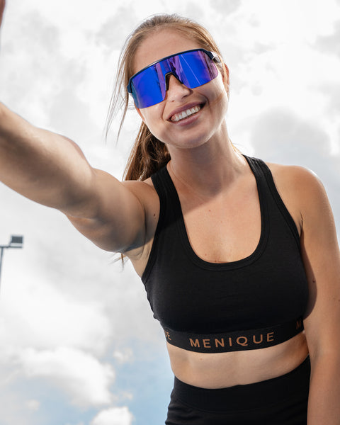 A close-up of a smiling woman wearing a black sports bra and blue reflective sunglasses, reaching one arm out toward the camera against a cloudy sky.