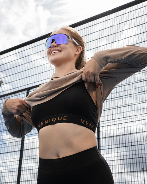 A woman outdoors by a wire fence wearing a black sports bra and black bottoms, adjusting a sheer tan long-sleeved crop top over her shoulders while wearing blue reflective sunglasses.