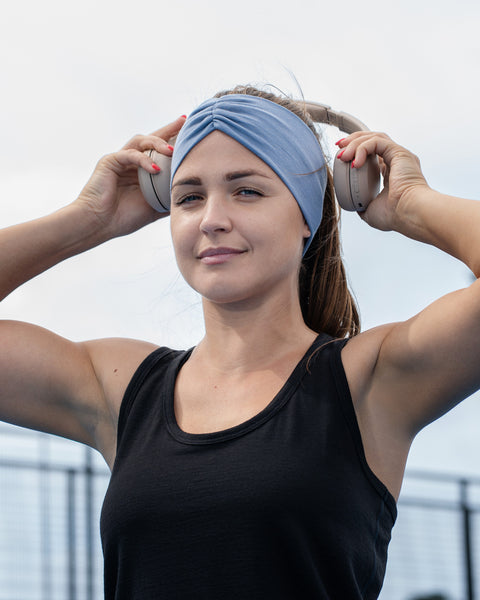 A close-up of a woman outdoors wearing a black tank top and a wide light blue headband, holding white headphones over her ears with both hands.