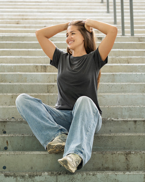 A smiling woman sitting cross-legged on concrete outdoor stairs, wearing a dark grey t-shirt and loose blue jeans, with both hands resting behind her head.