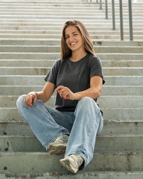 A smiling woman sitting cross-legged on concrete stairs, wearing a dark short-sleeved t-shirt and loose light blue jeans.