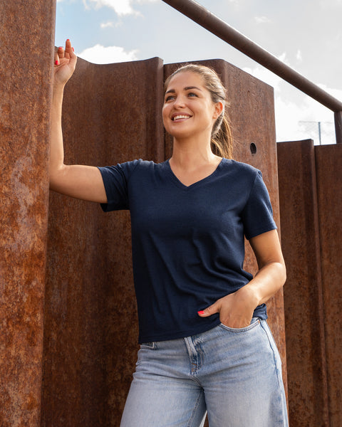 A smiling woman leaning against a rust-colored corrugated metal wall, wearing a dark blue v-neck t-shirt and blue jeans.