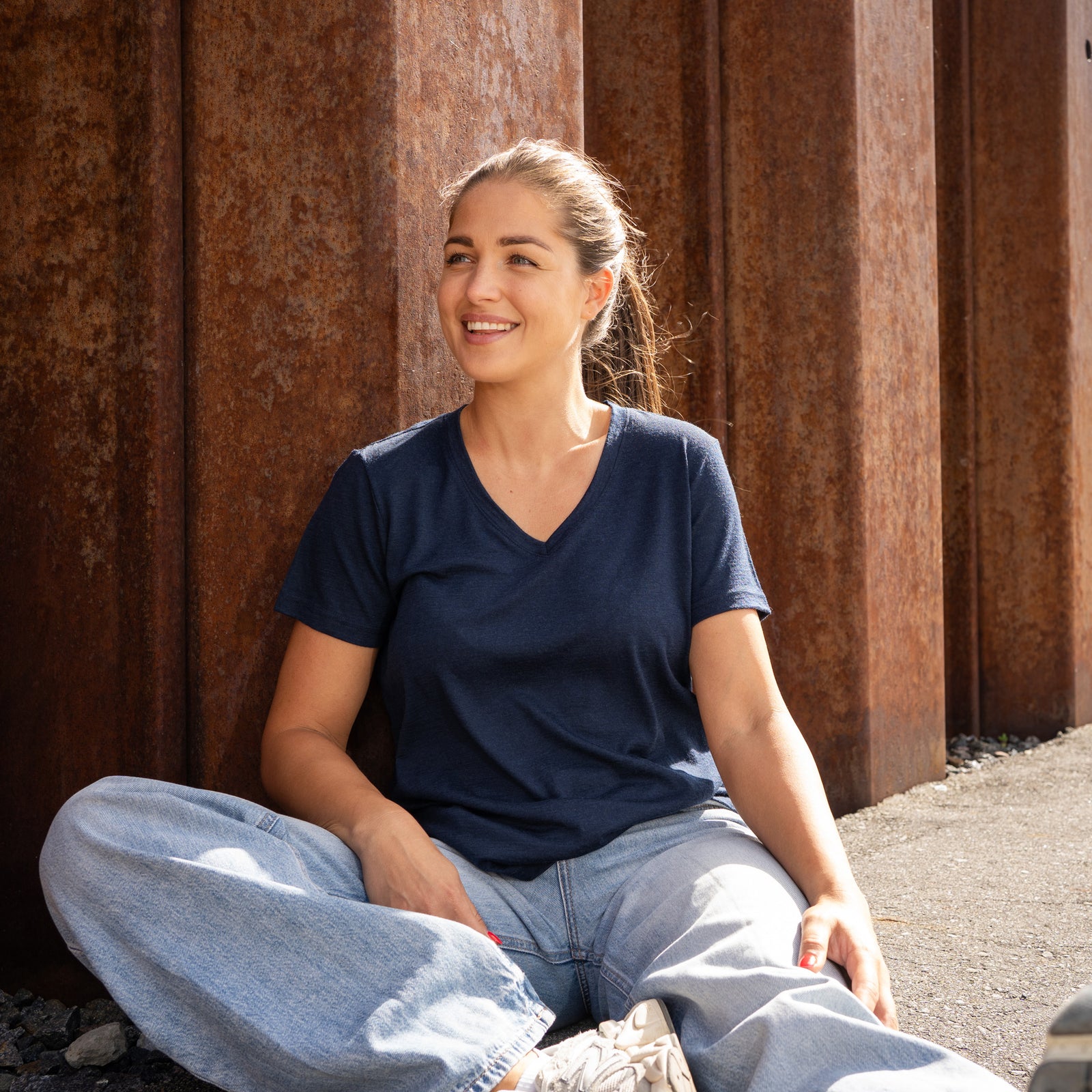 A woman standing next to a textured, rusty metal wall, smiling and looking away from the camera. She is wearing a navy blue v-neck t-shirt and blue jeans, with her right hand resting gently on the metal structure and her left hand in her pocket.