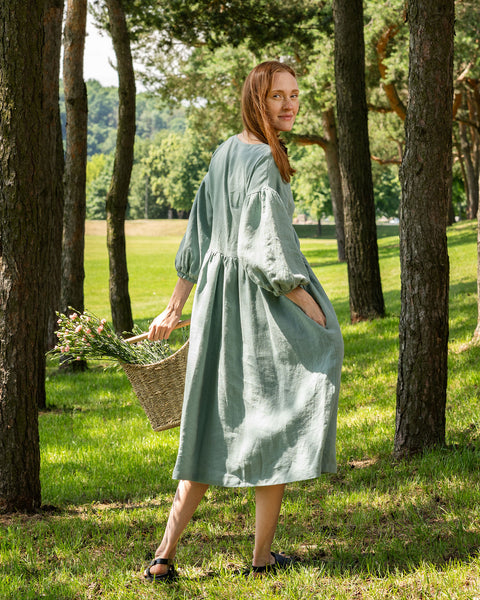 A person with reddish-brown hair stands in a grassy, wooded area, looking back over their shoulder towards the camera. They are wearing a mint green linen dress with puffed sleeves and black sandals. They are holding a woven basket filled with flowers in their left hand.