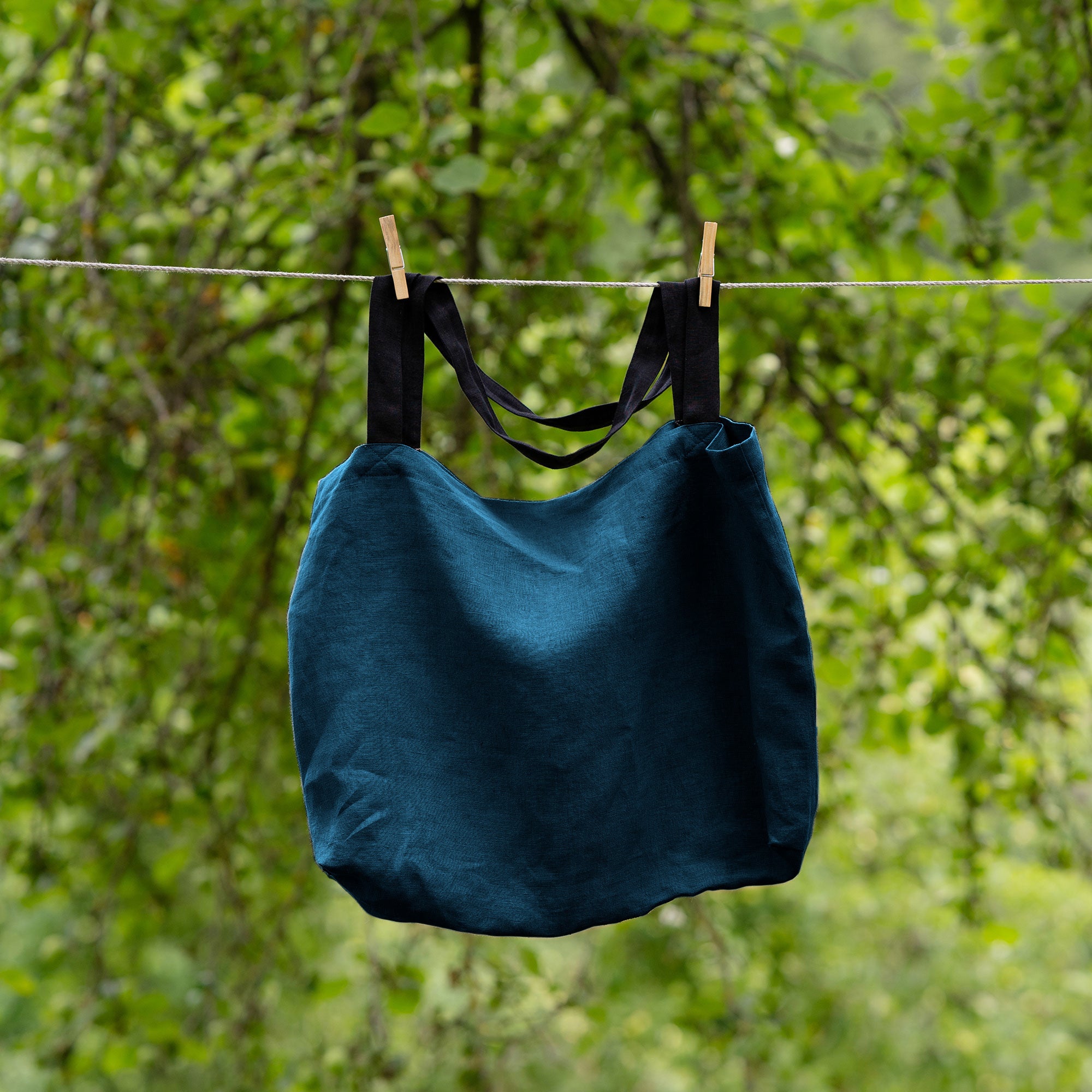 The menique linen tote bag in stone green color with black handles hung on a clothesline outside.