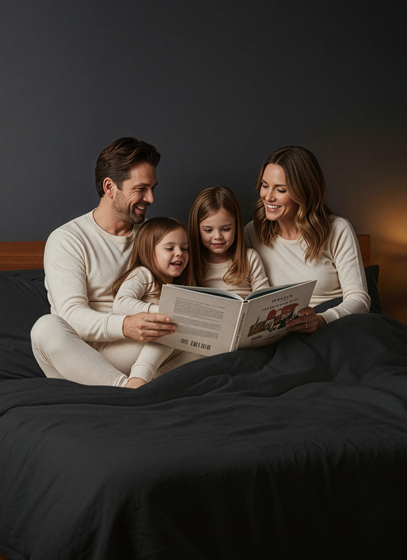 A family of four sits together in bed, smiling as the adults read a book to the two young children