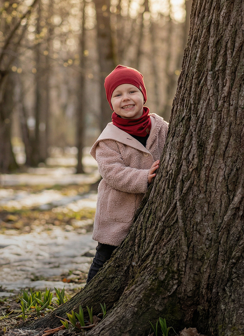 Child in a pink coat and red hat standing next to a tree in a forest
