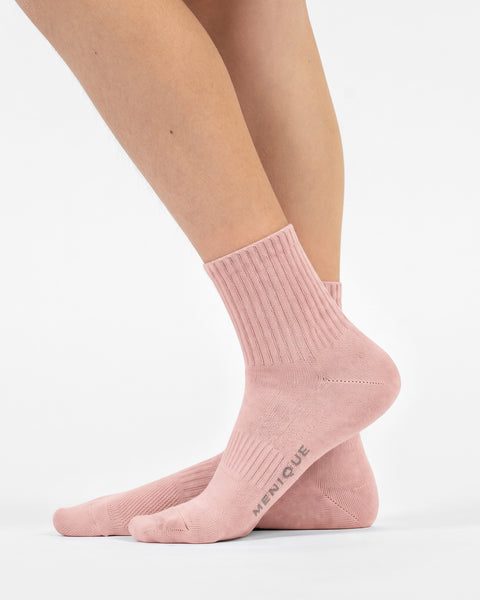 A side-profile view of a person's feet wearing light pink crew socks, revealing the word "MENIQUE" printed on the sole of the right foot.