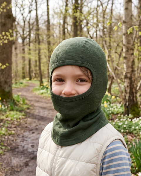 A close-up portrait of a young child smiling gently while standing on a dirt path in a forest. The child is wearing a dark olive green knitted balaclava that covers their head, neck, and lower face, leaving only their eyes and nose visible. They are also wearing a light cream-colored quilted vest over a blue and white striped long-sleeved shirt. The softly blurred background features bare trees, patches of green foliage, and small early spring flowers.