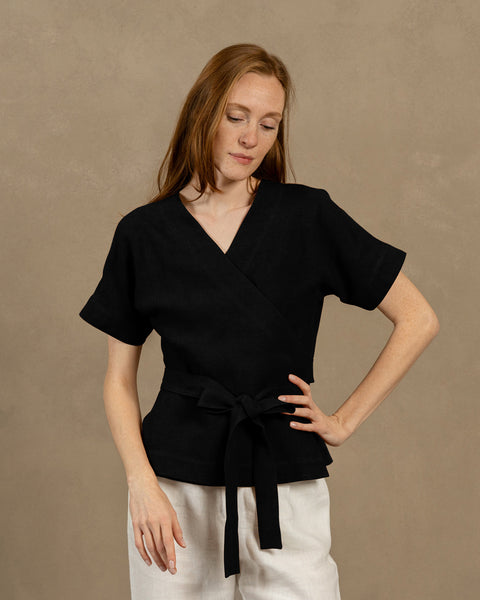 A fair-skinned person with long, auburn hair, wearing black V-neck linen wrap top with short, wide sleeves and light-colored pants. They are holding the ends of the top&#39;s tie in front of them, forming a bow.