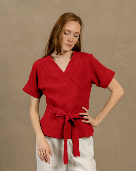 A fair-skinned person with long, auburn hair, wearing V-neck linen wrap top with short, wide sleeves and light-colored pants. They are holding the ends of the top&#39;s tie in front of them, forming a bow.