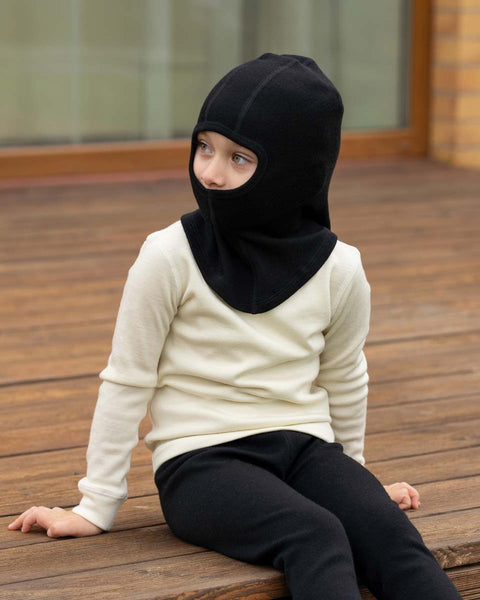 A young child wearing a black balaclava, a cream-colored long-sleeve shirt, and black pants sits on a wooden deck, looking away from the camera.