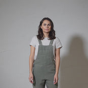 A woman with shoulder-length brown hair and blue eyes models stone green pinafore jumpsuit over a light-colored t-shirt against a textured gray backdrop in a short video.
