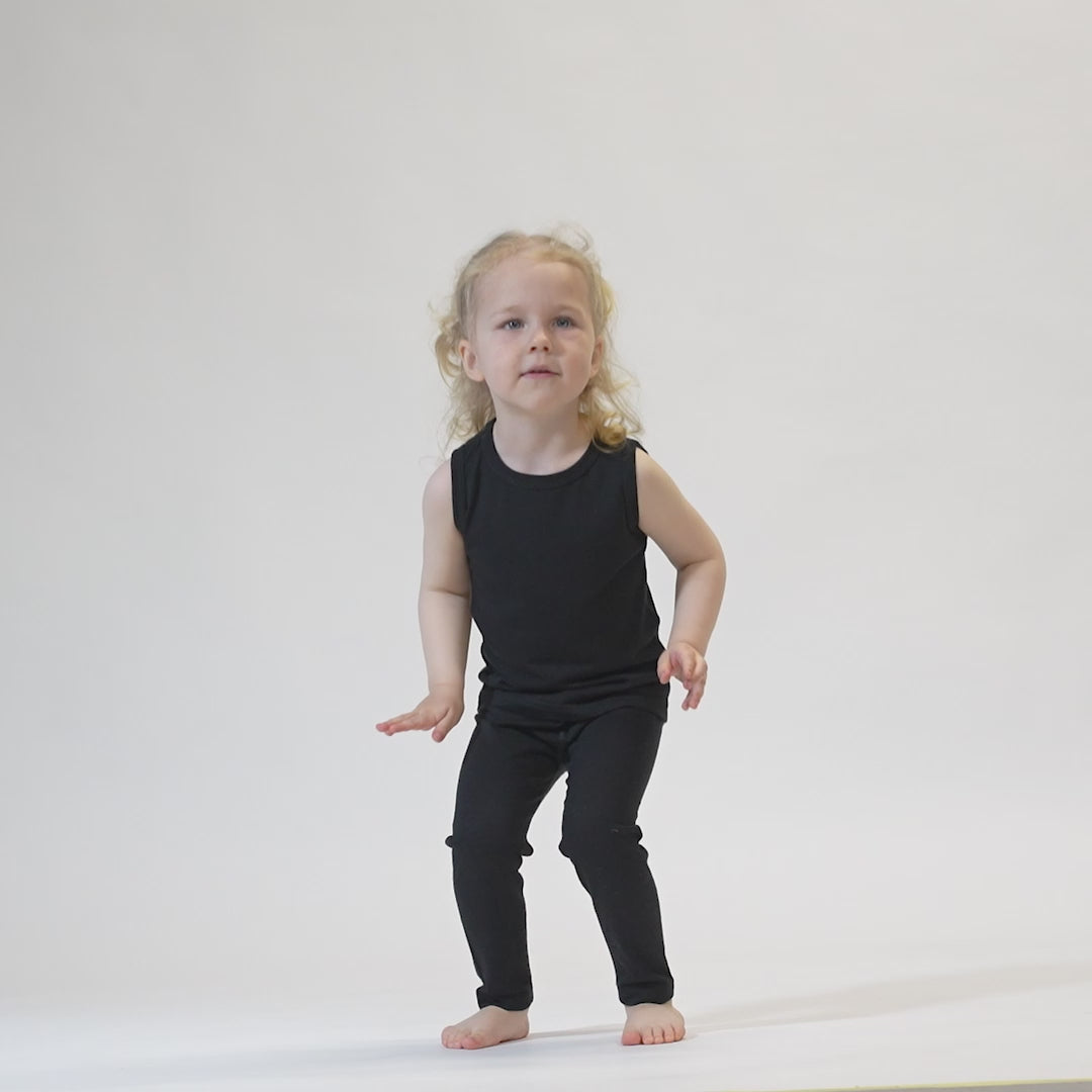 A young child with blonde hair, wearing a black menique top and black pants, stands against a white background with their arms raised above their head. The child is looking directly at the camera.