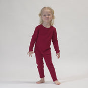 A young child with light, curly hair stands barefoot against a white background, wearing a long-sleeved, menique merino wool top and matching royal cherry pants. The child is looking at the camera with a slight smile and their hands are slightly open.