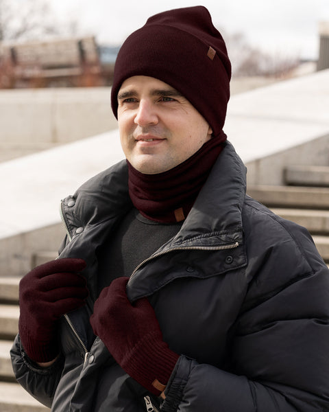 Man wearing a royal cherry Merino wool winter set including beanie, gloves, and neck warmer, styled with a black puffer jacket outdoors in cold weather.
