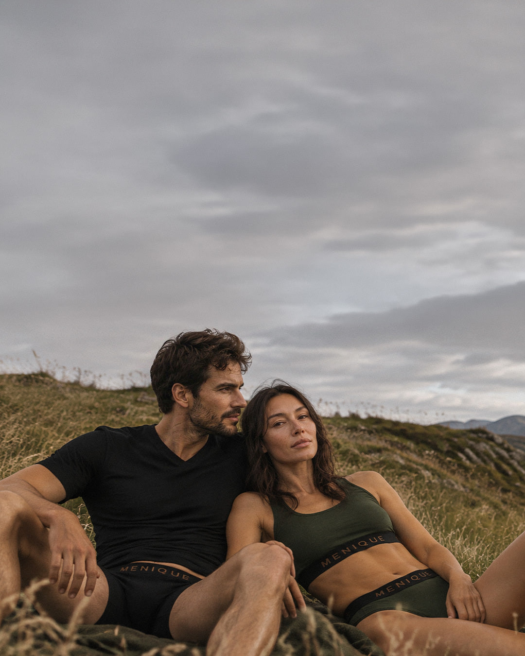 A man and a woman lounging in a grassy field near a lake, with mountains in the background under an overcast sky. The man is wearing a black v-neck t-shirt and black boxer briefs, while the woman wears an olive green sports bra and matching underwear. Both sets of underwear feature "MENIQUE" branded waistbands.