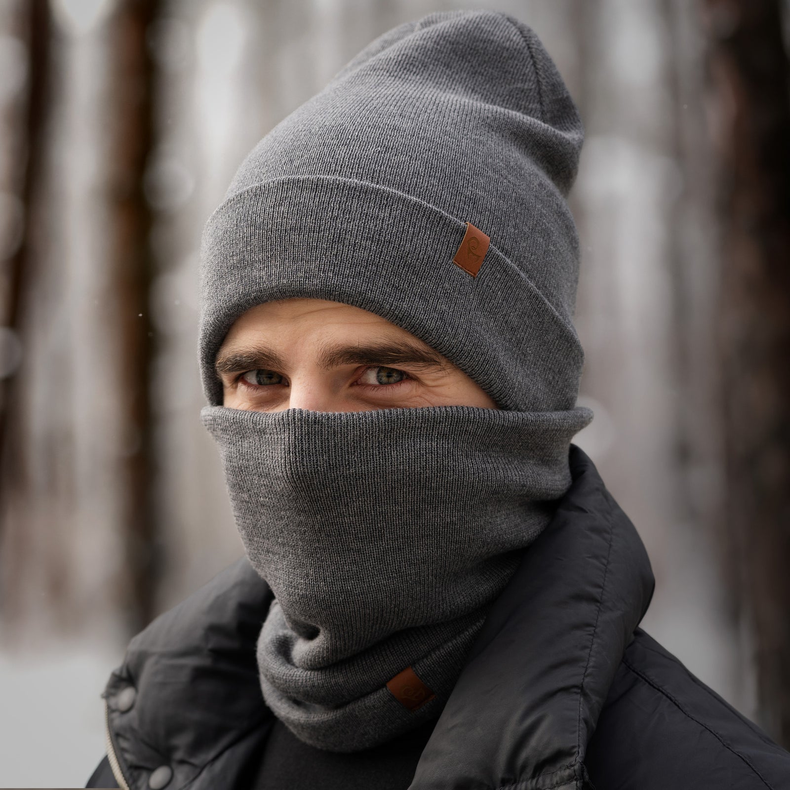 Man wearing grey merino beanie and neck gaiter, standing outdoors in snowy forest