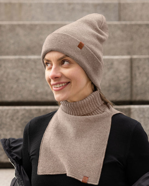 A woman with a fair complexion is pictured outdoors, smiling and looking off-camera to her right. She's wearing a light brown or taupe ribbed knit beanie with a small, square brown logo patch on the left side of the cuff. Around her neck, she has a matching knit neck warmer; this piece features a thick ribbed turtleneck and a rectangular, flat-knit bib that extends down her chest. This neck warmer also has a small brown logo patch at the bottom corner of the bib.