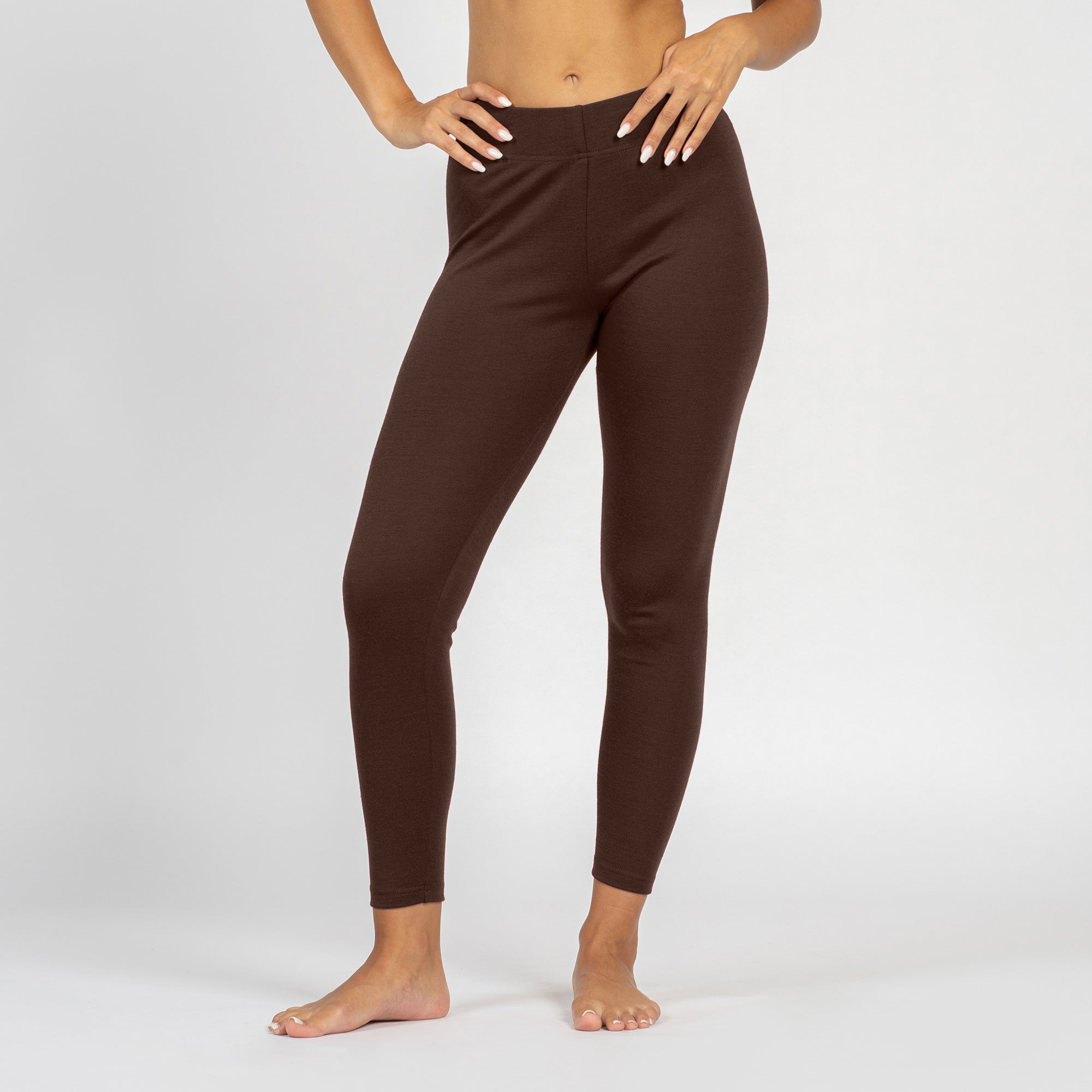 A person from the waist down stands in profile, facing left, against a plain white background. They are barefoot and wearing fitted, brown leggings, with their hands resting on their hips.