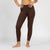 A person from the waist down stands in profile, facing left, against a plain white background. They are barefoot and wearing fitted, brown leggings, with their hands resting on their hips.