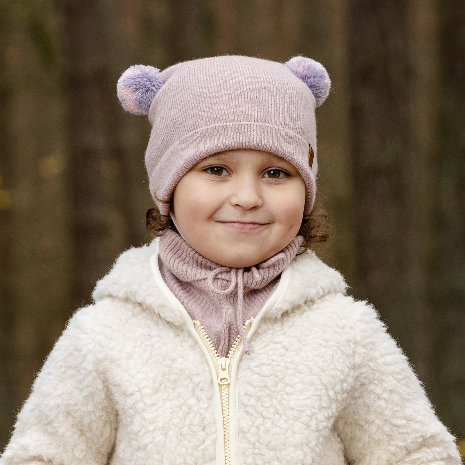 A young child with blue eyes and a slight smile, looks directly at the viewer. They are wearing a beige knit hat with brown pom-pom ears, a beige turtleneck or neck warmer, and a light pink sherpa-style coat. The background is a blurred outdoor scene with muted green and brown tones.