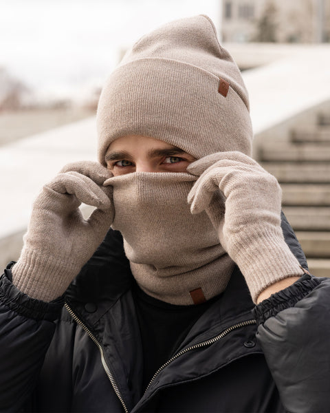 A man wearing a creamy beige knitted beanie and a matching neck gaiter pulls the gaiter up to cover most of his face, with only his eyes visible. He is also wearing creamy beige knitted gloves and a dark puffer jacket. The background is a blurred outdoor setting with stone steps.