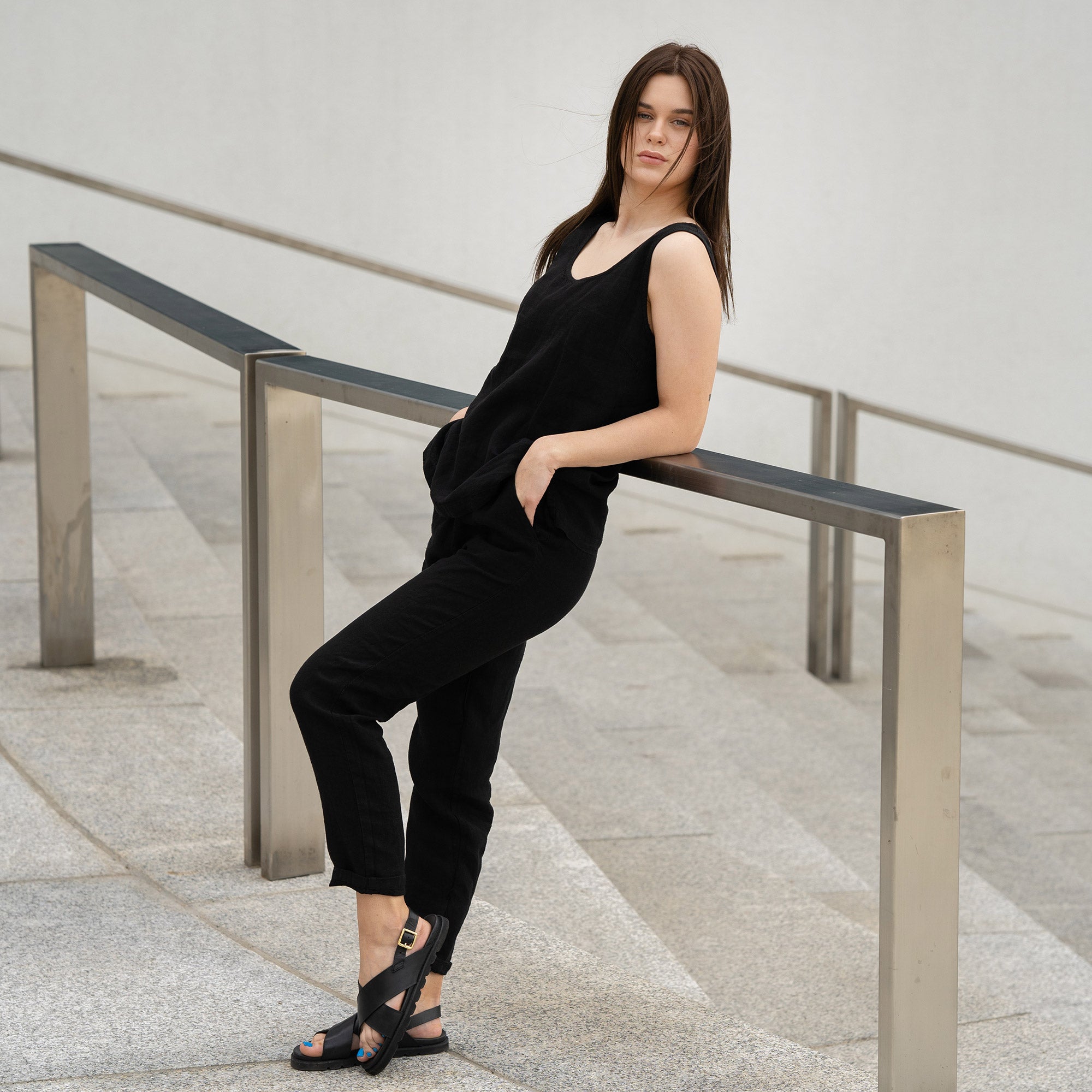A young girl posing outdoors in a city on a concrete stairs, wearing linen pure black tank top Hana, and pure black pants dakota. 