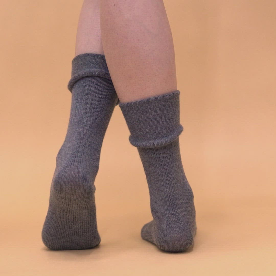 A woman wearing long merino wool socks in dark gray color, rolled down, turning around in the video.