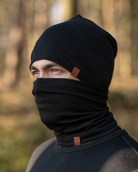 The Menique Men&#39;s Merino Beanie and Gaiter Set in Black color. The man is wearing the accessory set in a forest, in fall time.